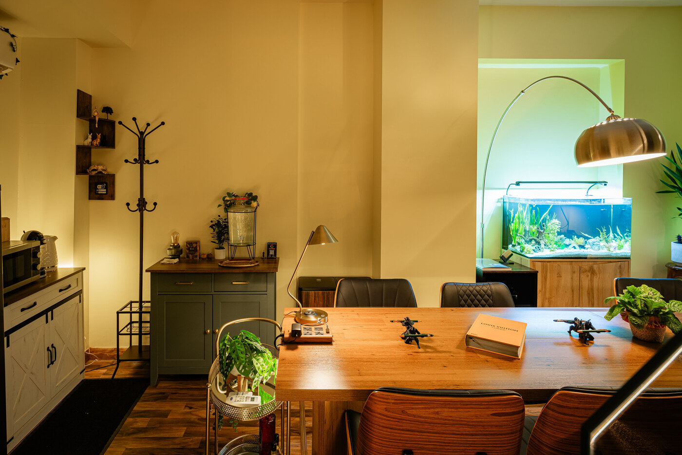 BC Atelier by the hour — the warmly lit long dining table with the brass arc lamp, plants, shelves and the studio fish tank